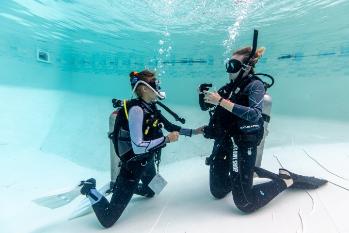 Two scuba divers underwater in a pool, practicing hand signals.