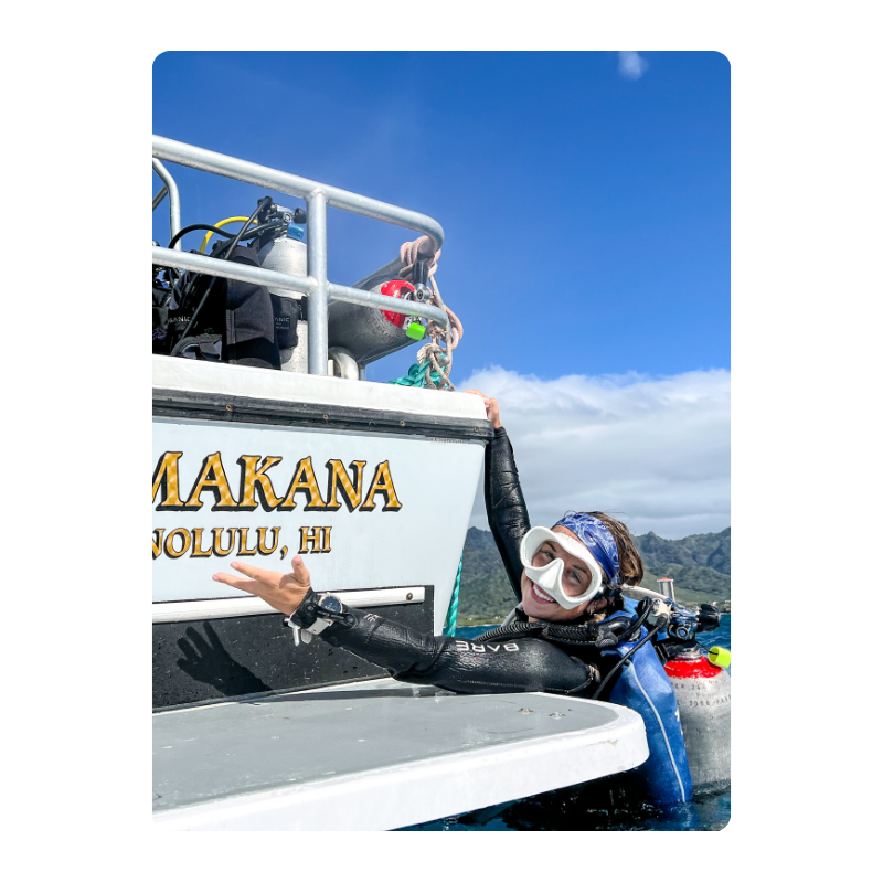 Diver in wetsuit smiles, holding onto a boat named Makana in Honolulu, HI.