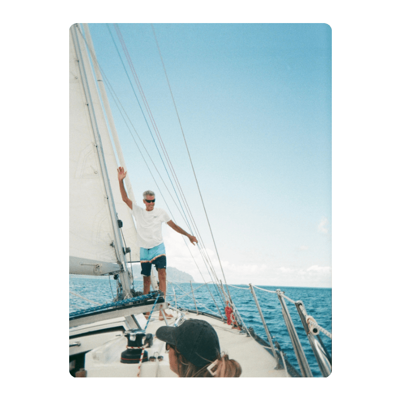 Two people on a sailboat, one standing and waving, sunny day at sea.