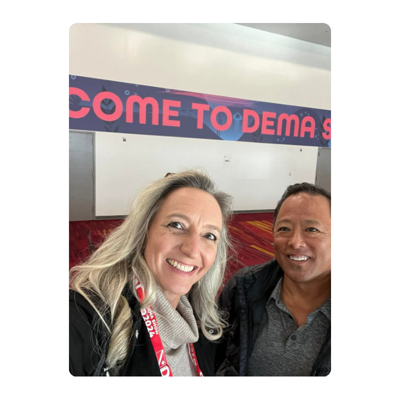 Two people smiling in front of a sign that reads 'WELCOME TO DEMA SHOW'.