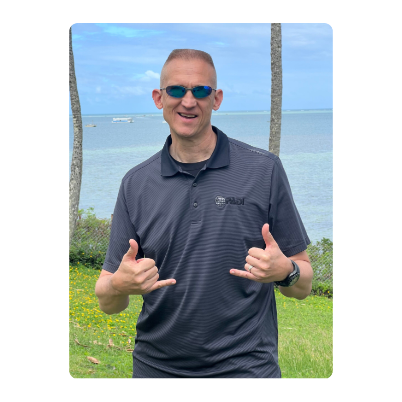 Person in sunglasses makes shaka sign near ocean, wearing dark polo shirt.