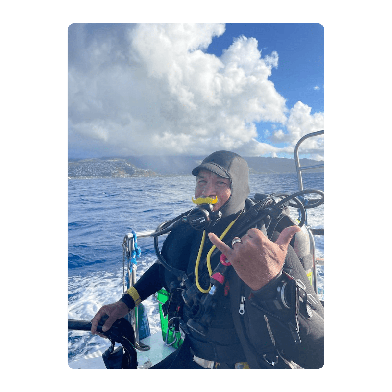 Scuba diver on a boat, smiling and making a shaka sign, with ocean and cloudy sky in background.