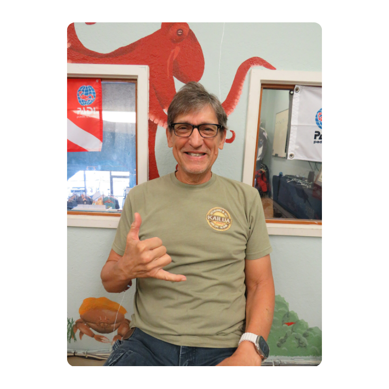 Man seated, smiling, making shaka sign, with octopus mural and diving signs in background.