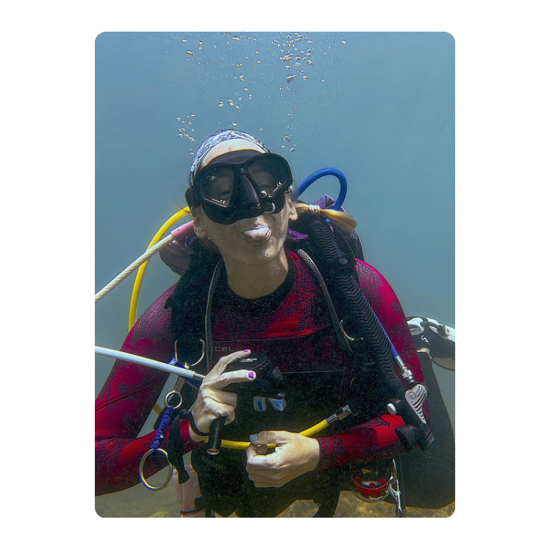 Scuba diver underwater, wearing black goggles and red wetsuit, bubbles rising to the surface.