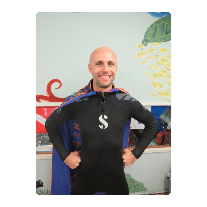 Man in wetsuit with cape poses in front of colorful aquatic mural.