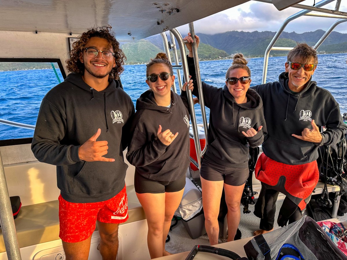 Four people in matching hoodies pose on a boat with ocean and mountains in the background.