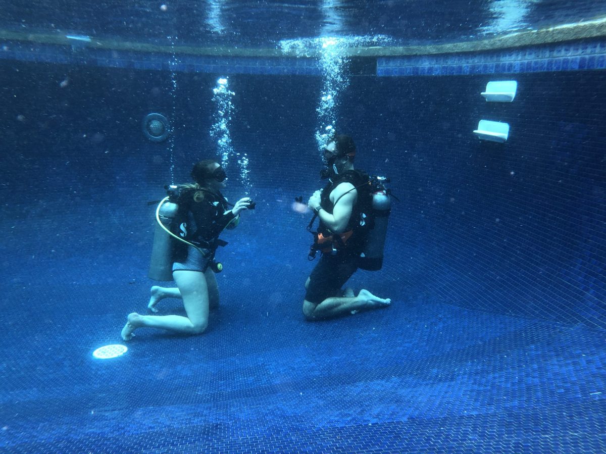 Two scuba divers kneeling underwater in a pool, facing each other.