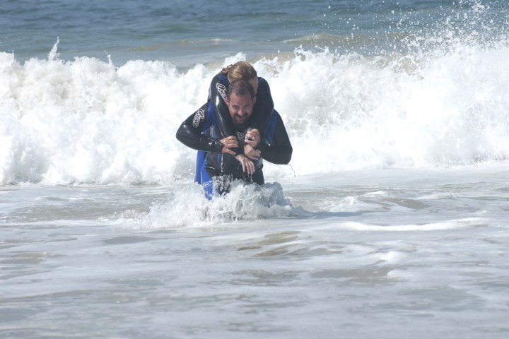 Two people in wetsuits in ocean waves, one carrying the other on their back.
