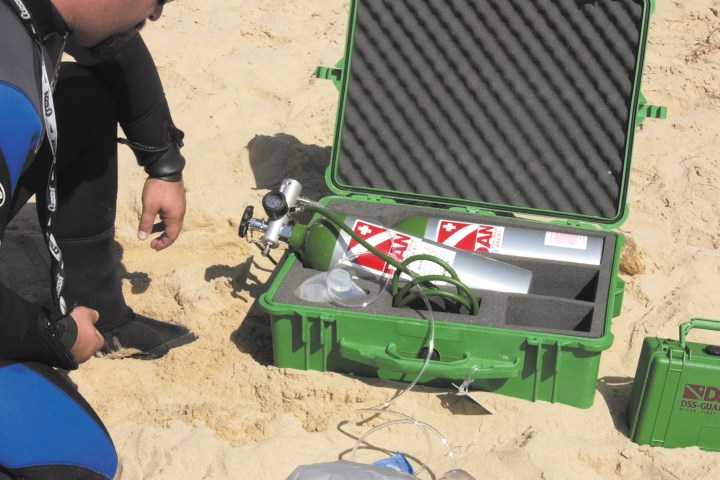 Person lying on beach with oxygen mask, another in wetsuit kneeling nearby, and a green open medical box.