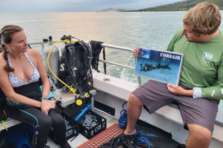 Two scuba divers on a boat, one holding a sign about the Corsair.